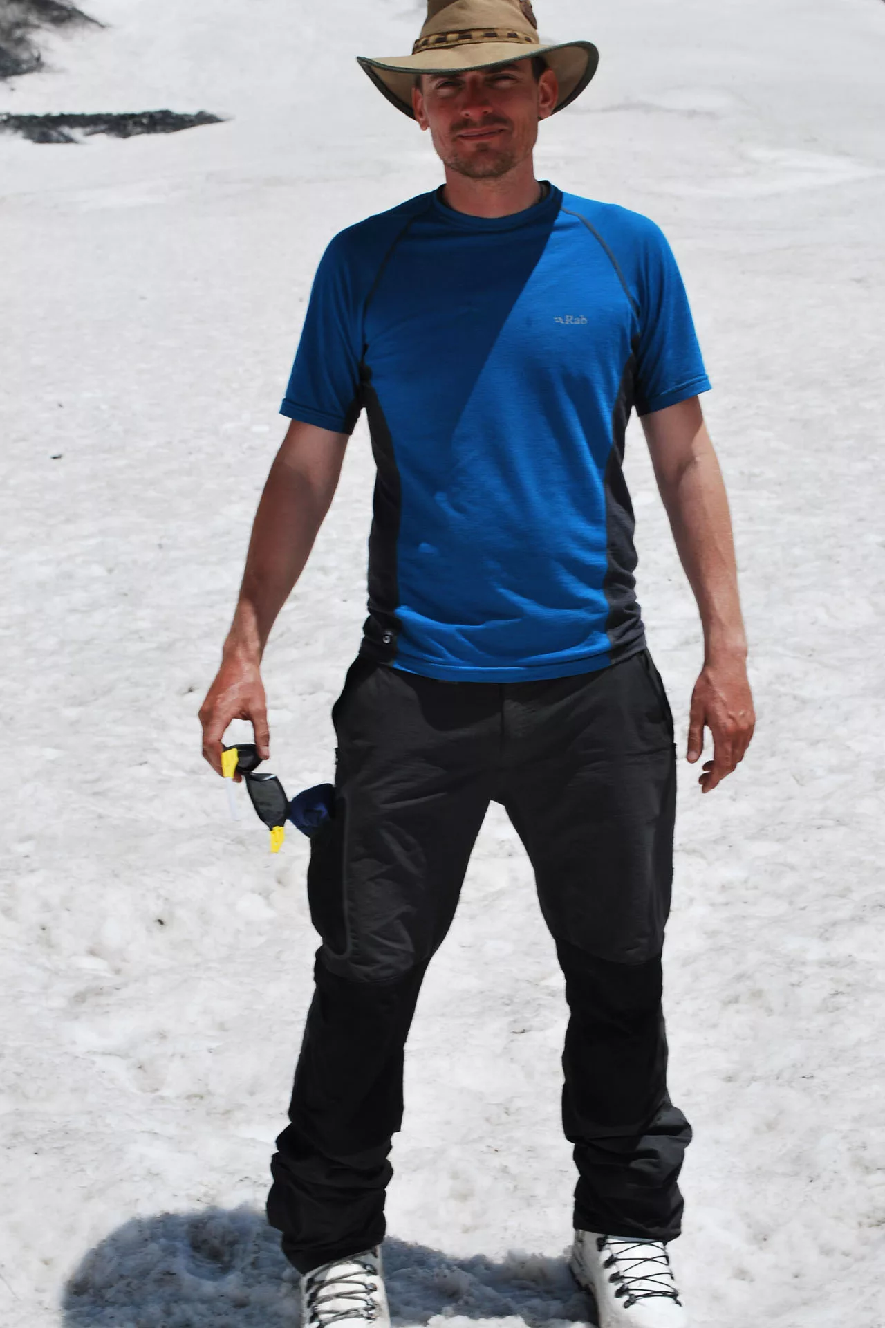 Client standing on a glacier in a blue rab tshirt, black pants, a sun hat, googles in his right hand, and mountain climbing boots