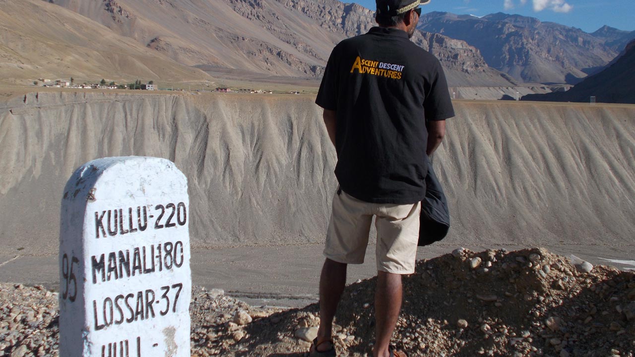 Mountain guide wearing an ascent descent adventures logo t-shirt stands next to a milestone indicating the distance from Spiti to Manali