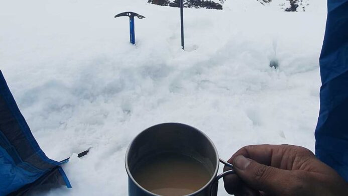 Climber's view from blue tent vestibule, holding a cup of tea with an ice axe stuck in the snow in front, surrounded by snowy mountain landscape.